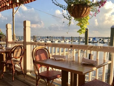 Waterfront restaurant view with outdoor seating overlooking marina at sunset, wicker chairs, and red-striped awning.