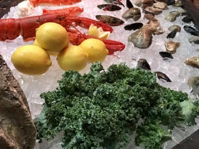 Fresh seafood platter with shrimp, oysters, clams, mussels, lobster, lemons, and kale on ice at a gourmet buffet.