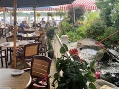 Outdoor restaurant patio with tables, chairs, and lush garden by a small waterfall, under a striped canopy near the sea shore.