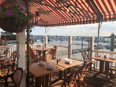 Outdoor dining area at seaside restaurant with striped canopy, overlooking marina with boats, offering a scenic view.